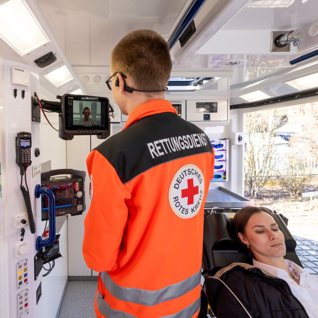 Vorführung Telenotarzt: Mann vom Rettungsdienst kommuniziert mit einem anderen über kleinen Bildschirm, rechts eine Frau auf Liege