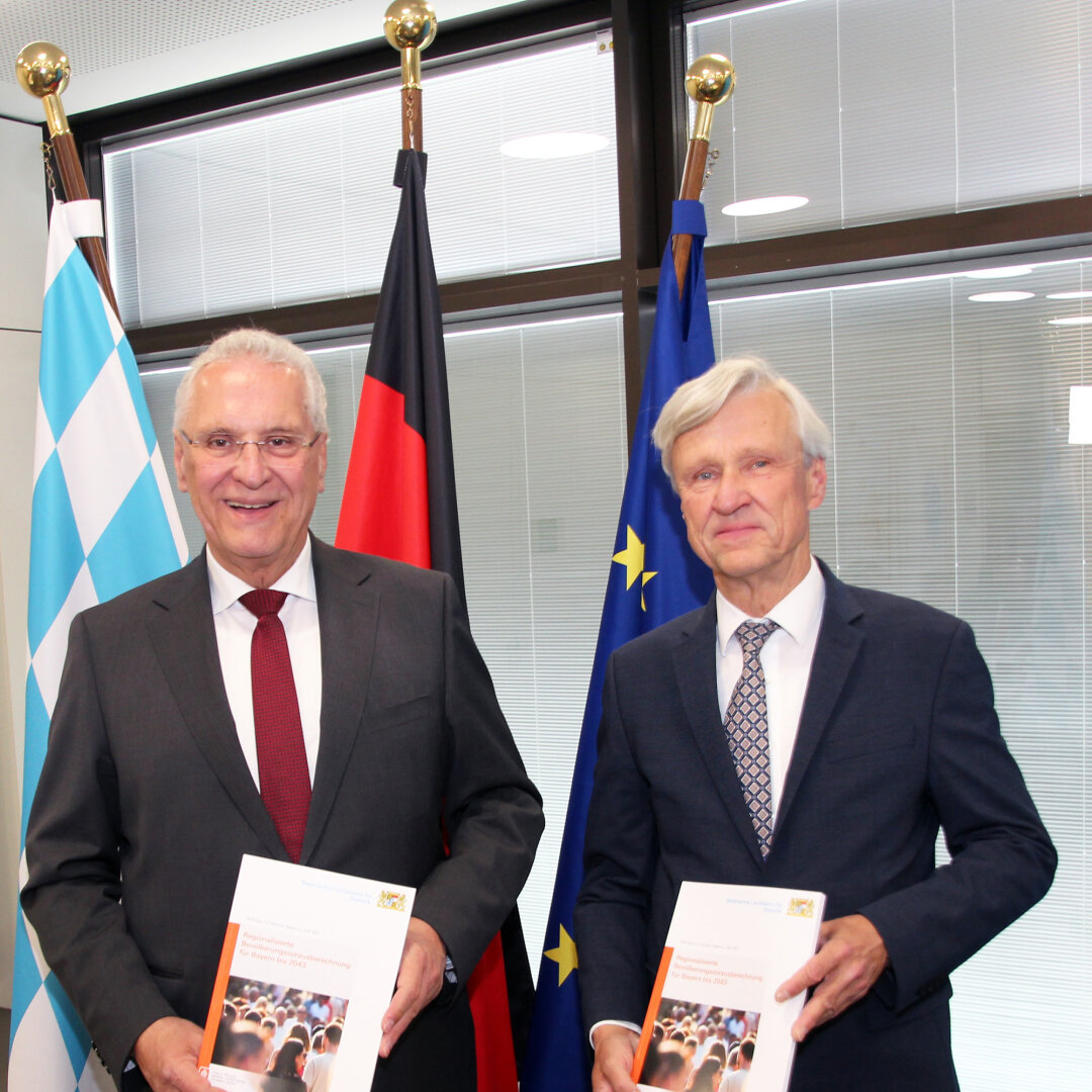 links Innenminister Joachim Herrmann, links Dr. Thomas Gößl, Amtsleiter Bayerisches Landesamt für Statistik stehen vor einem Fahnenset und halten die gebundene Ausgabe Bevölkerungsvorausberechnung 2043 in den Händen