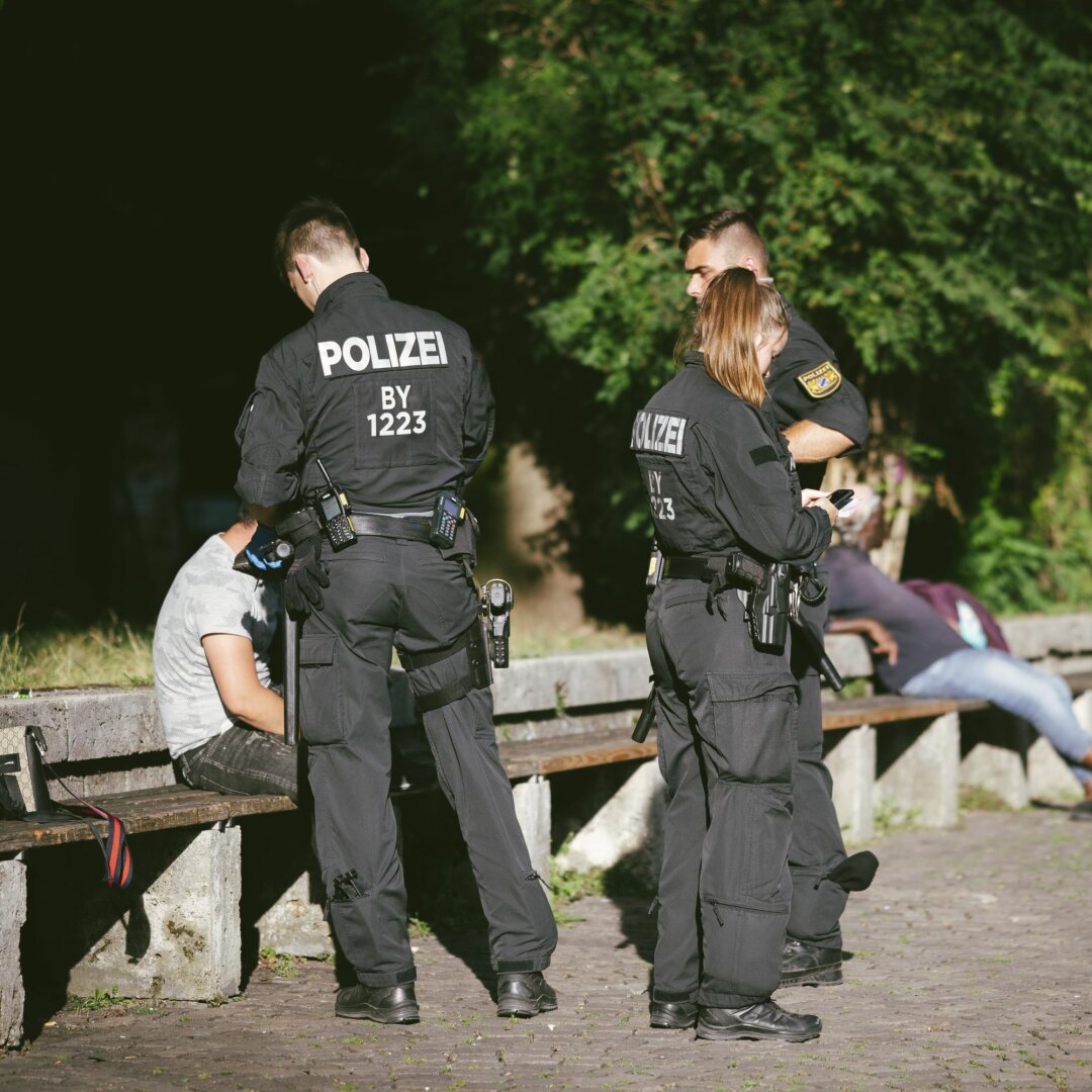 Auf dem Bild sind eine Polizistin und zwei ihrer Kollegen zu sehen, wie sie im Alten Botanischen Garten in München eine auf einer Bank sitzende Person kontrollieren.