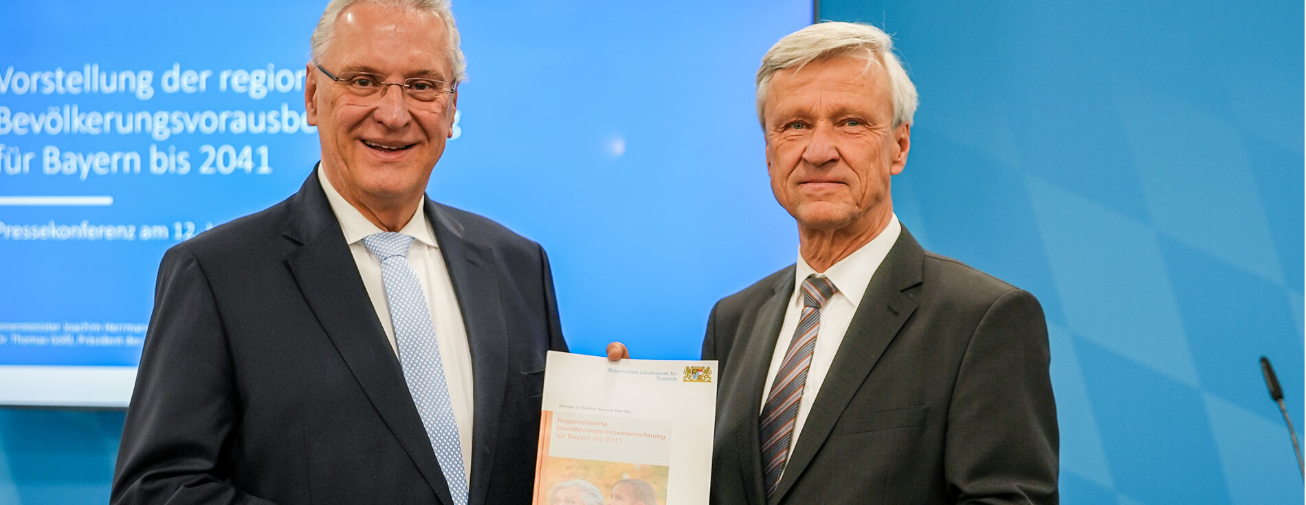 Innenminister Joachim Herrmann und Dr. Thomas Gößl mit Broschüre in der Hand, im Hintergrund Präsentation