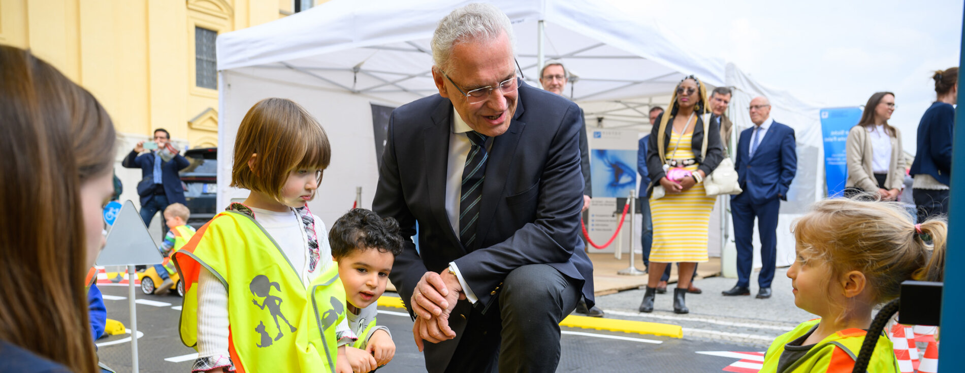 Innenminister Joachim Herrmann kniet zu Kindern in Verkehrswarnfesten auf Bobycars