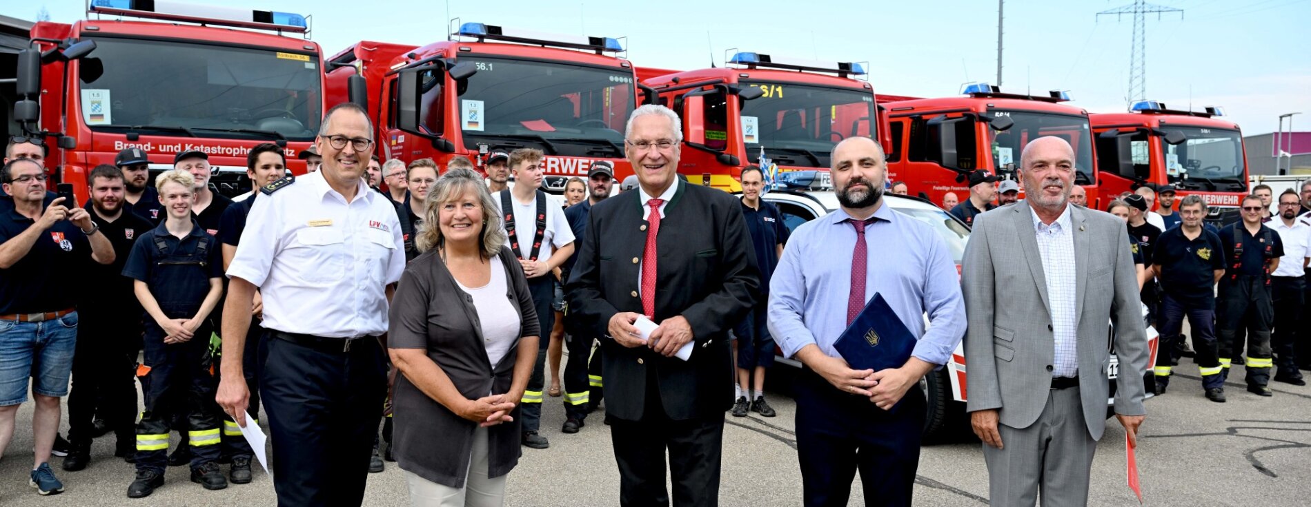 Innenminister Joachim Herrmann, ukrainischer Generalkonsul Oleksii Kravets und der Vorsitzender des Landesfeuerwehrverbandes Bayern (LFV) Johann Eitzenberger mit weiteren vor einer Gruppe Helferinnen und Helfern und Feuerwehrfahrzeuge