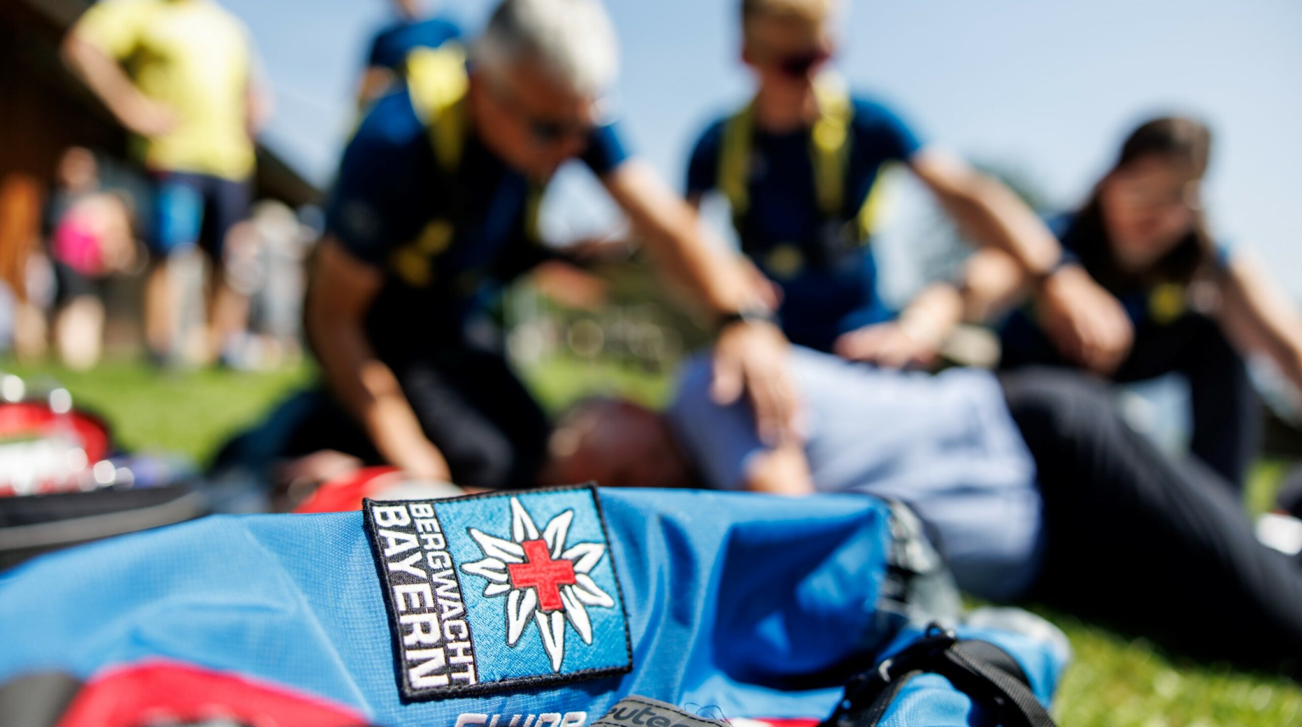 Auf dem Foto ist im Vordergrund ein Rucksack mit dem Logo der Bergwacht Bayern zu sehen und im Hintergrund sind zwei Bergwacht-Mitglieder bei Erste-Hilfe-Übungen verschwommen erkennbar.