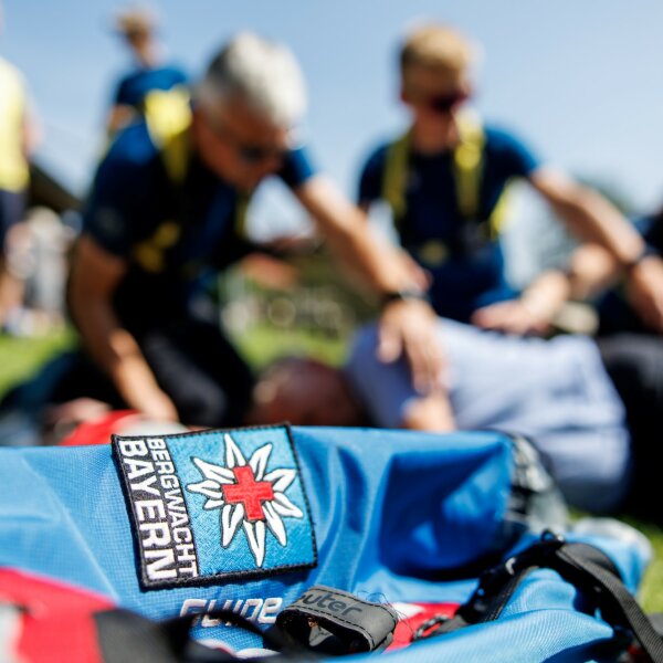 Auf dem Foto ist im Vordergrund ein Rucksack mit dem Logo der Bergwacht Bayern zu sehen und im Hintergrund sind zwei Bergwacht-Mitglieder bei Erste-Hilfe-Übungen verschwommen erkennbar.