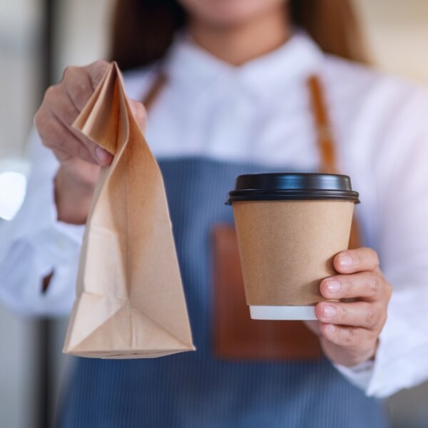 Frau hält einen Coffee-to-go-Becher und eine Papiertüte in der Hand