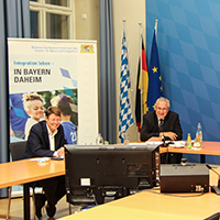 Innenminister Herrmann und Mitarbeiterinnen des StMI im Sitzungssaal vor einem Bildschirm
