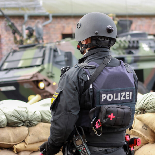 Bewaffneter Polizist von hinten hinter Berg von Säcken, im Hintergrund Panzerfahrzeug