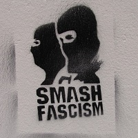 Das Bild zeigt ein an eine Wand gesprühtes Graffiti. Dieses Graffiti in schwarzer Farbe zeigt zwei vermummte Gesichter und den Text „SMASH FASCISM“